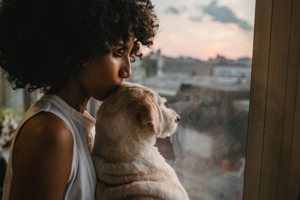 Crop African American female holding and kissing adorable puppy while standing near window with closed eyes