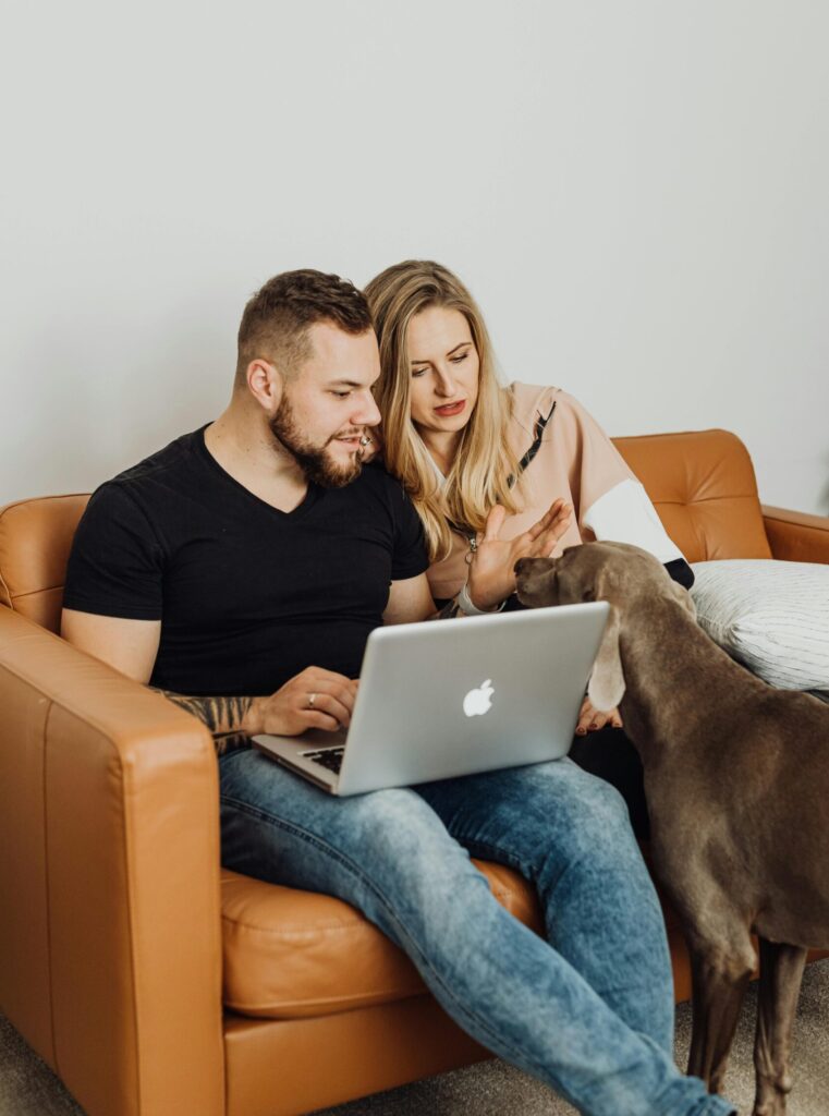 Couple lounging on a sofa with laptop and dog, indoors. Caucasian adults engaging with technology and pet.